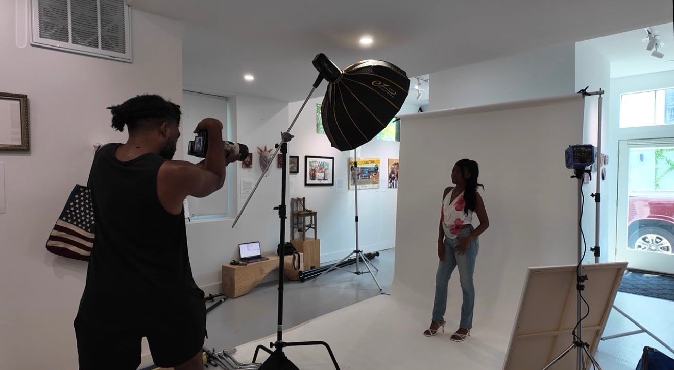 A lifestyle scene inside a studio space showing a photographer taking a photo of a woman posing against a white seamless backdrop. The studio is equipped with a large softbox, a second light stand, and an easel.